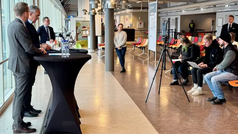 Två män står vid talarstolar inför närvarande från press och media under en pressträff