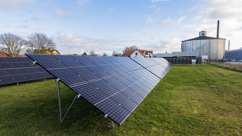 Solpaneler på ett grönt fält med industribyggnad och hus i bakgrunden