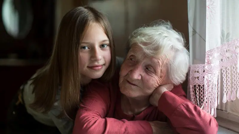 Flicka omfamnar äldre kvinna som sitter vid fönstret