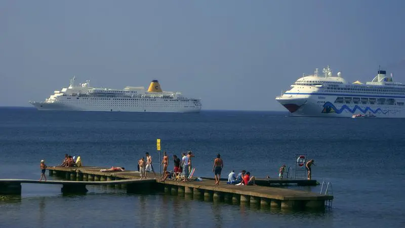 Två stora kryssnyngsfärjor på havet, badbrygga med människor som solar och badar i förgrunden