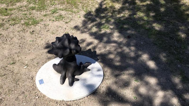 Ett litet svart träd av metall står i skuggan av ett stort levande träd
