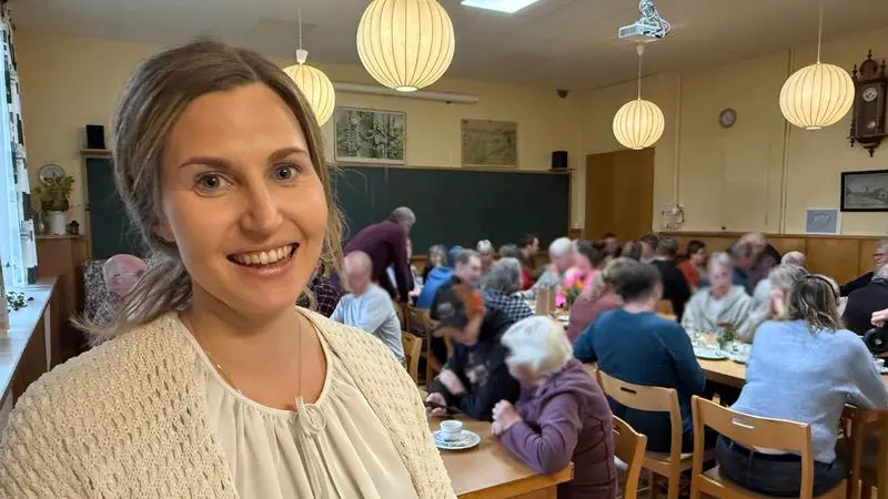 En glad kvinna står i en lokal där det sitter många människor och fikar.