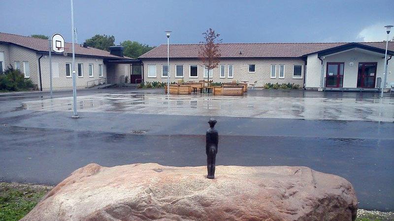 En liten figur i metall står på en stor sten, i bakgrunden syns en skolgård och två byggnader