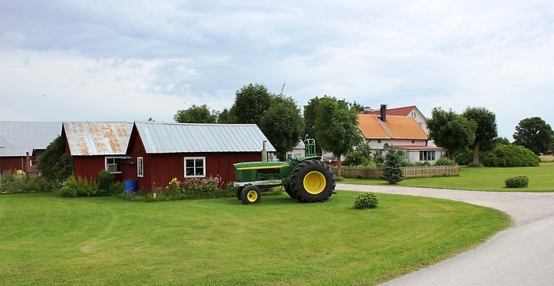 På vänster sida av vägen står en grön traktor, och bakom den finns flera röda stugor.