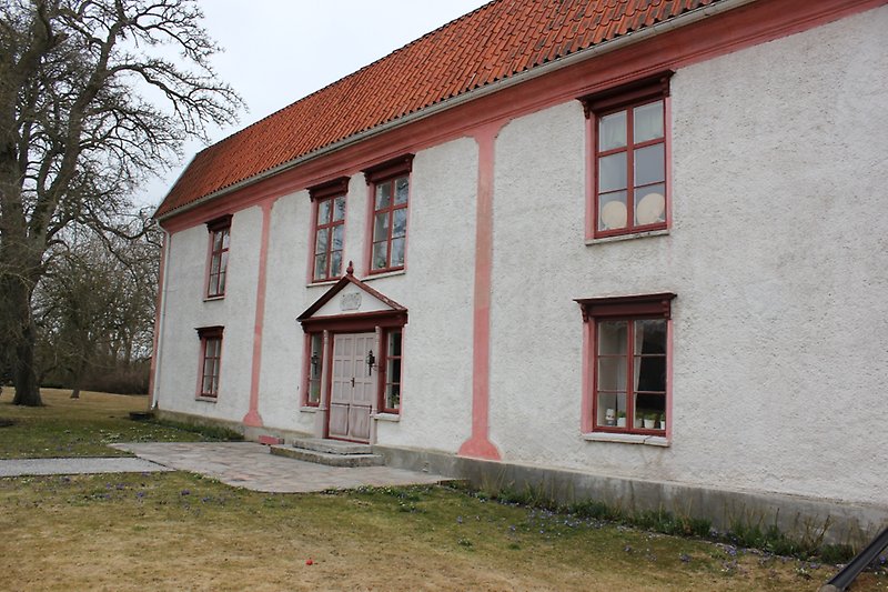 Ett vitt hus med rosa detaljer, en vit ytterdörr, röda fönster och ett rött tak.
