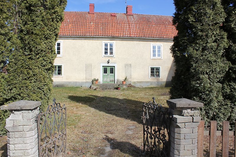 Bakom öppna grindar finns det ett beige hus med gröna och vita fönster och ett rött tegeltak.