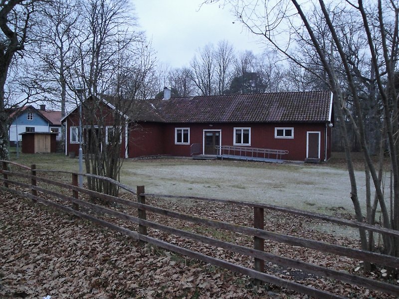 En röd envånings träbyggnad med vita fönster, bruna dörrar och ett rött tak med en skorsten.