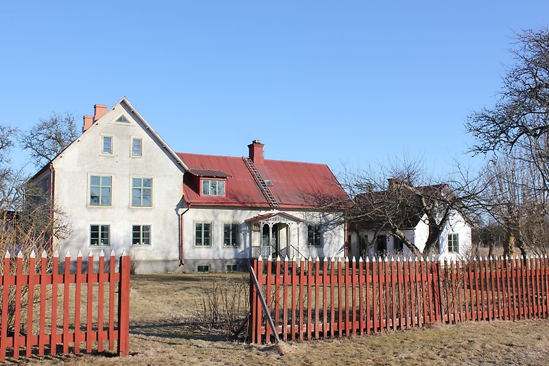 Bakom den röda staketet finns det ett vitt hus med gröna fönster och ett rött plåttak.