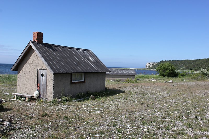 En liten fiskebod som ligger nära havet och har putsade väggar, ett svart plåttak med en skorsten.