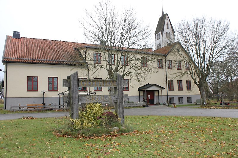 En gul tvåvåningsbyggnad med röda fönster och ett rött sadeltak med flera skorstenar. Höger om byggnaden ser man en kyrka.