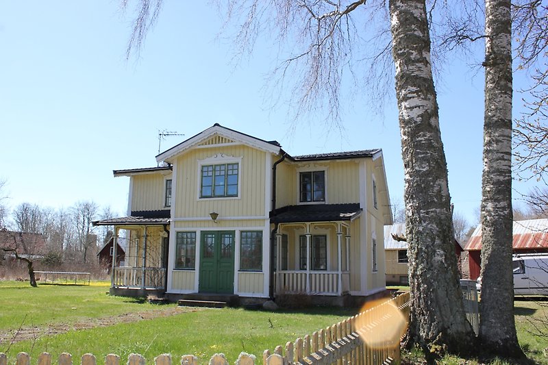 Ett ljusgult hus med en träfasad, gröna fönster, gröna dörrar och ett svart tak.