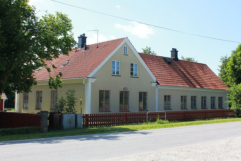 Ett ljusgult hus med röda fönster, ett rött tak och ett rött staket som ligger längs bilvägen.