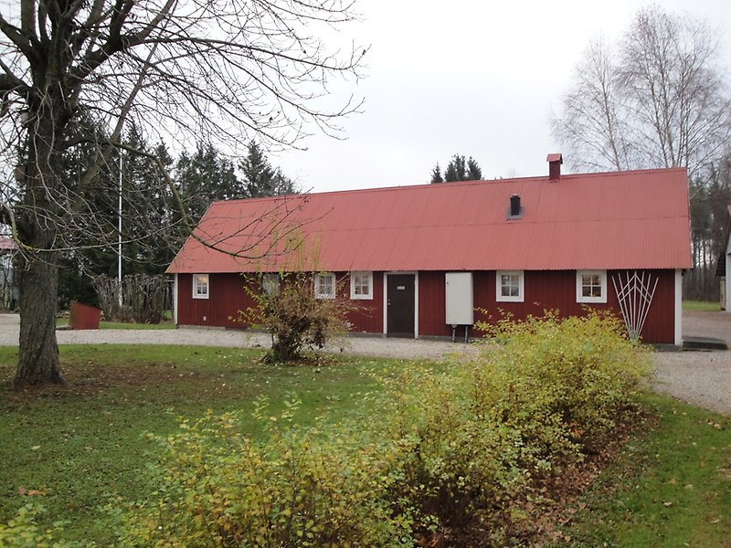 En röd byggnad med ett rött plåttak, vita fönster och en svart ytterdörr.