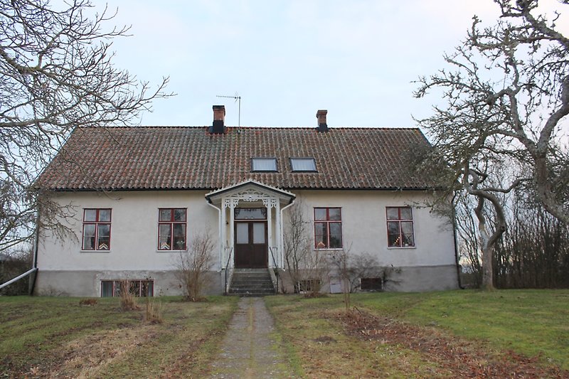 En vit envåningsbyggnad med rödvita fönster, en brun ytterdörr och ett sadeltak med två skorstenar.