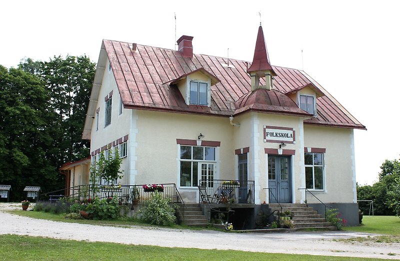 En ljusgul byggnad med vita fönster, blåa ytterdörrar och ett rött plåttak.