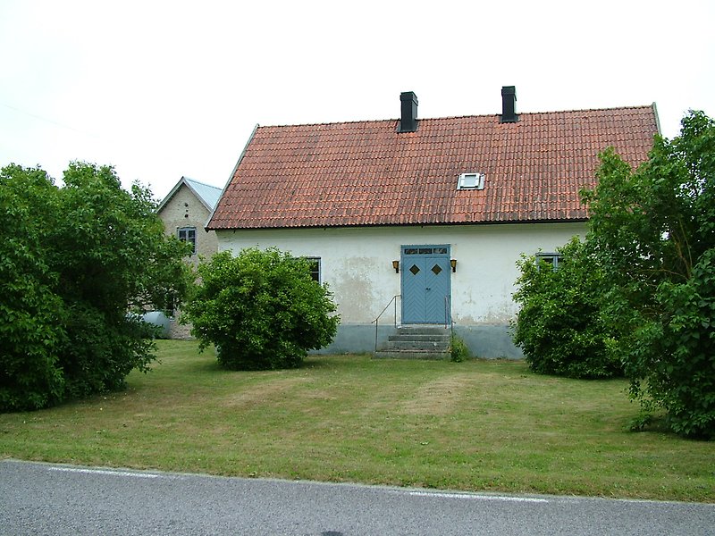 Längs bilvägen ligger ett vitt hus med blåa fönster, en blå ytterdörr och ett rött tak med två skorstenar.